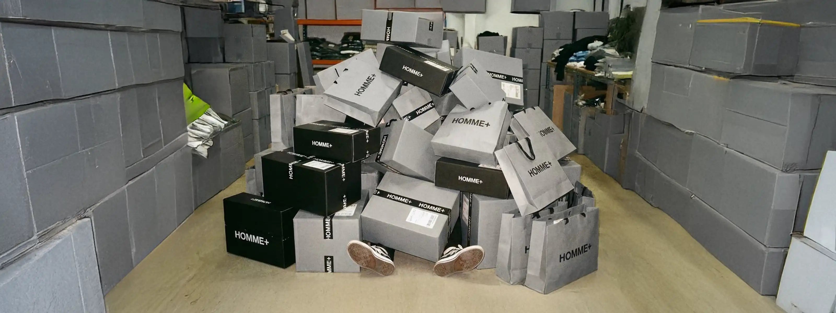 Black Friday sale banner for HOMME+ with a man covered by black and grey shipping boxes and shopping bags showing many online orders of menswear.