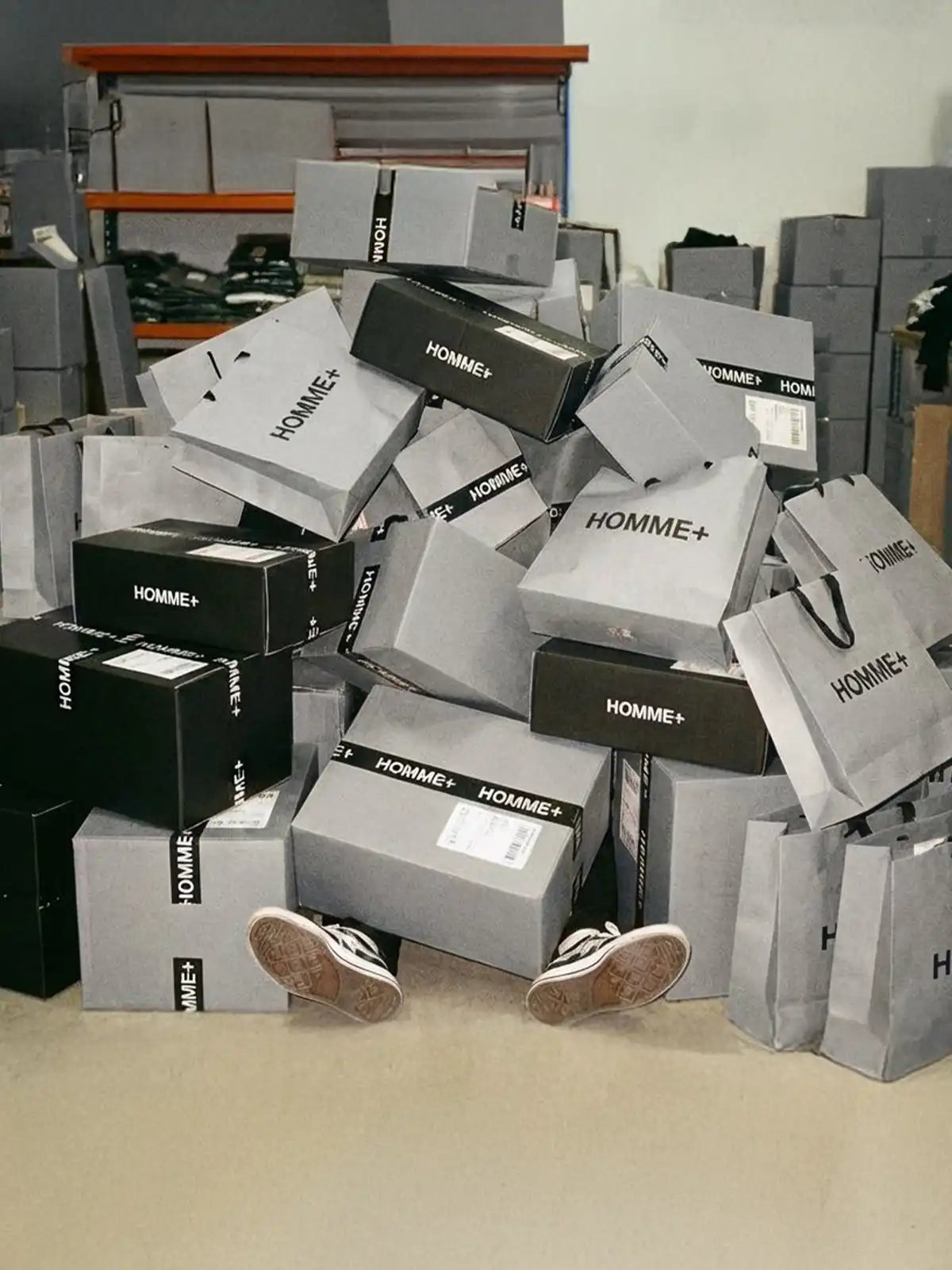 Black Friday menswear sale banner for HOMME+ with black and grey shipping boxes and shopping bags showing many online clothing orders.