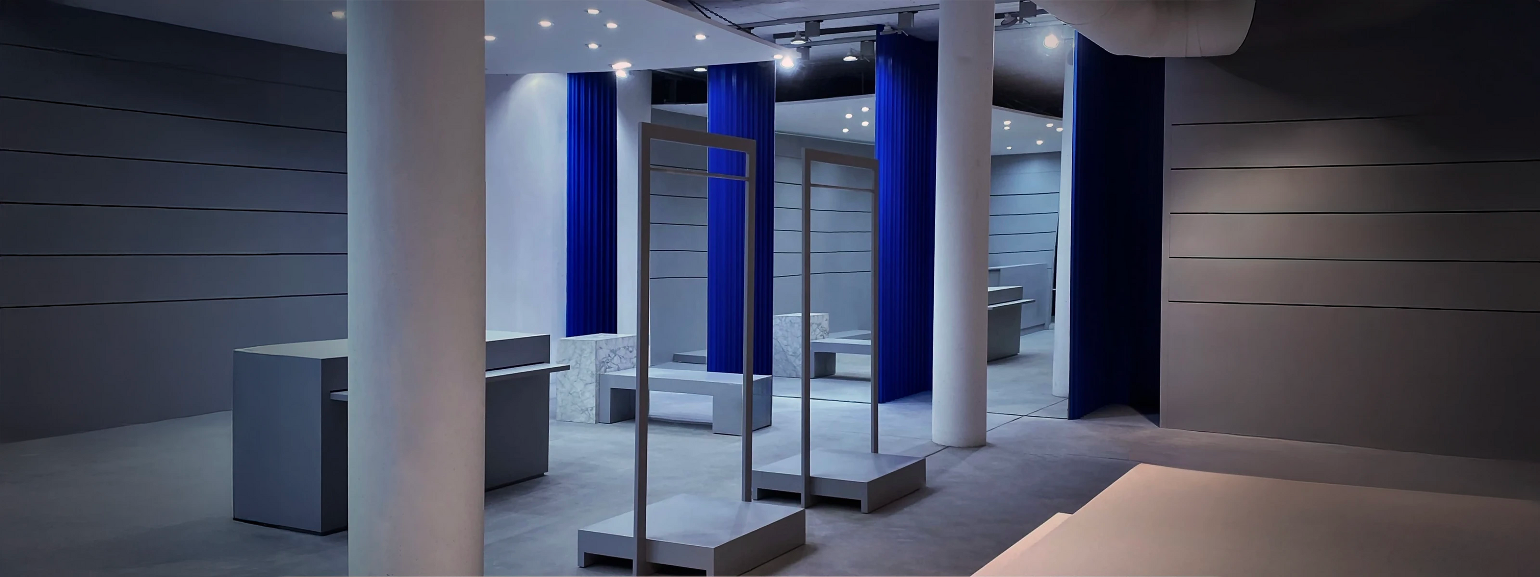 Interior of the HOMME+ fashion boutique in Ibiza, featuring a minimalist avant-garde design. The space is characterized by large white cylindrical columns, horizontal grey wall paneling, and dramatic floor-to-ceiling cobalt blue velvet curtains that mark the fitting room areas.