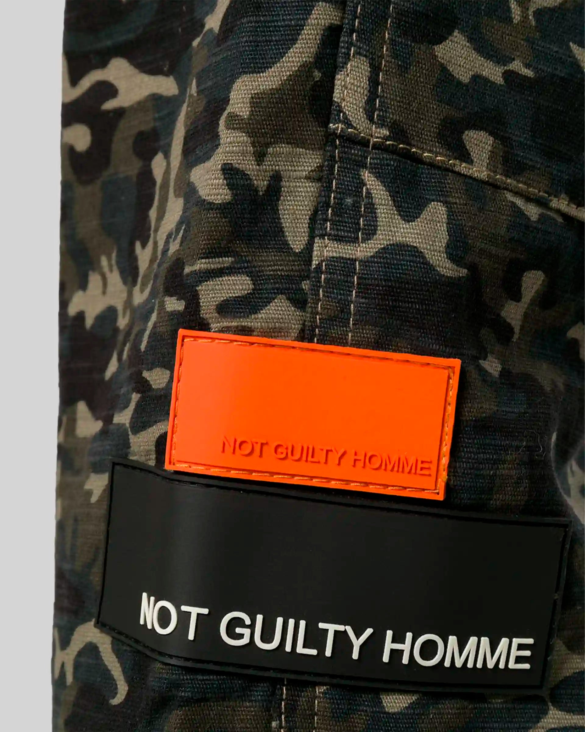 Close‑up of contrast Not Guilty Homme logo patches on camo fabric.