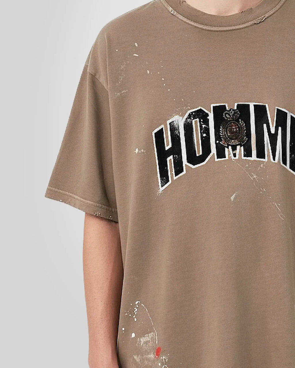 Close‑up of the tonal embroidered patch with distressed textured fill and the hand‑made paint stains on the beige t‑shirt.