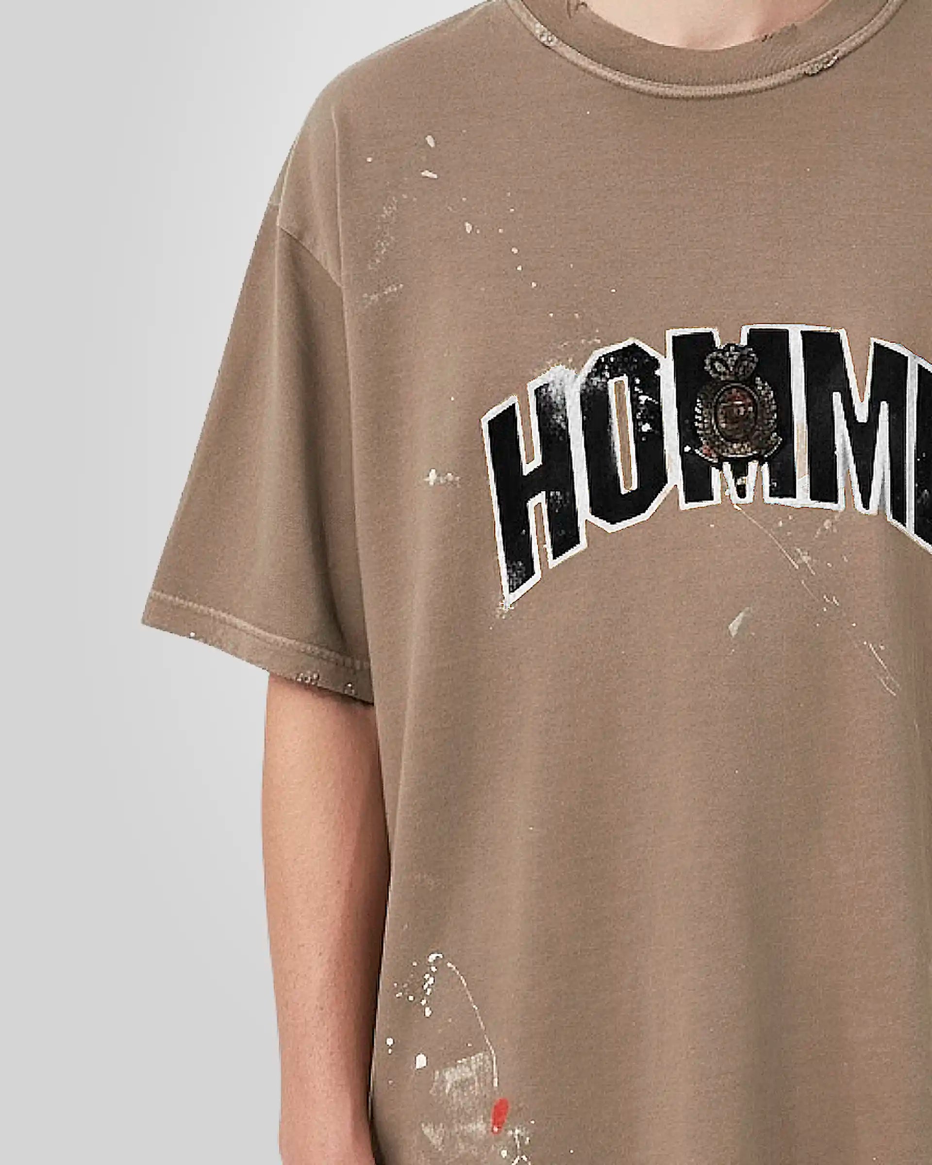 Close‑up of the tonal embroidered patch with distressed textured fill and the hand‑made paint stains on the beige t‑shirt.