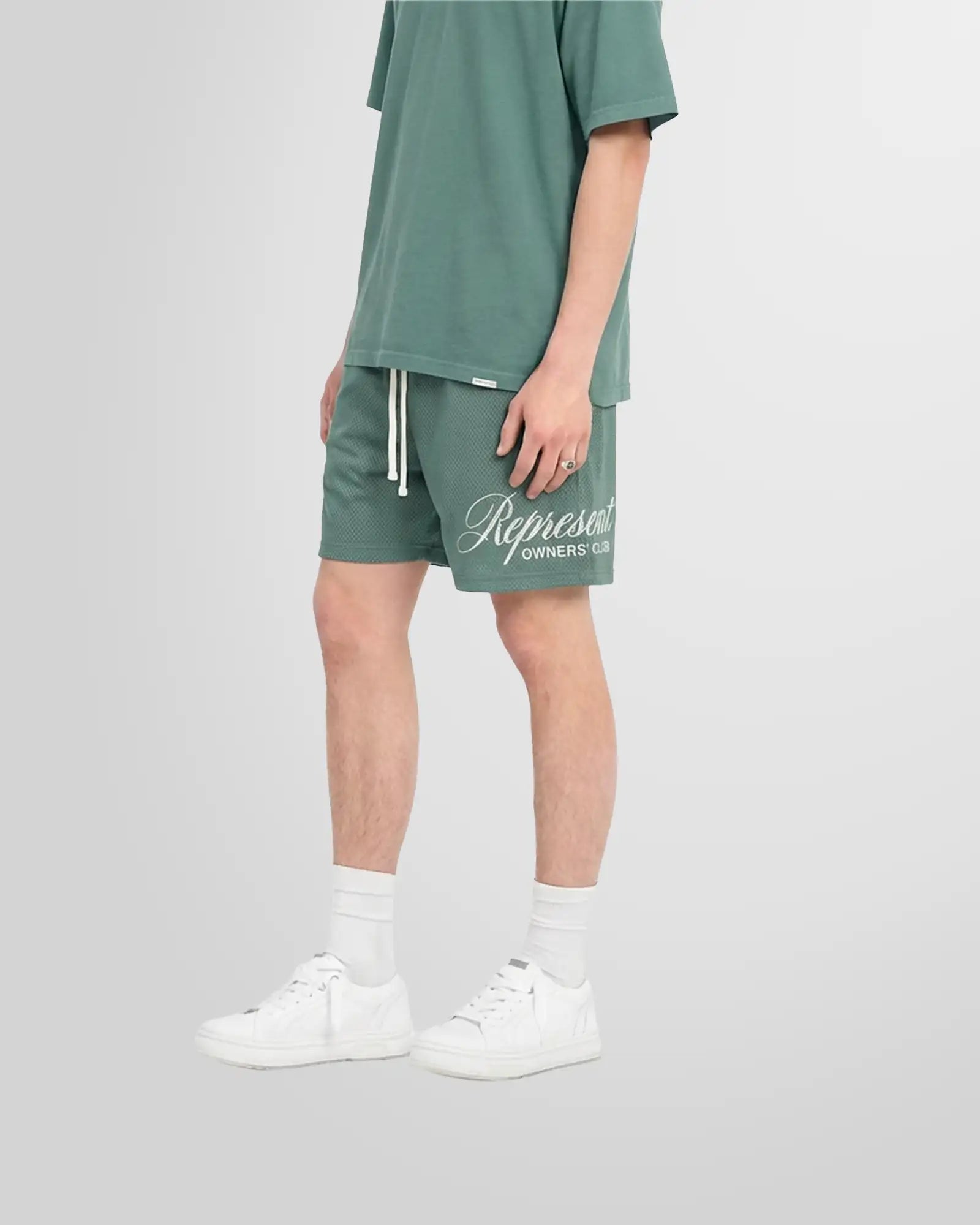 View from the side of a model wearing the Represent Owners’ Club Script Mesh Shorts in Vintage Green showing the print in white.