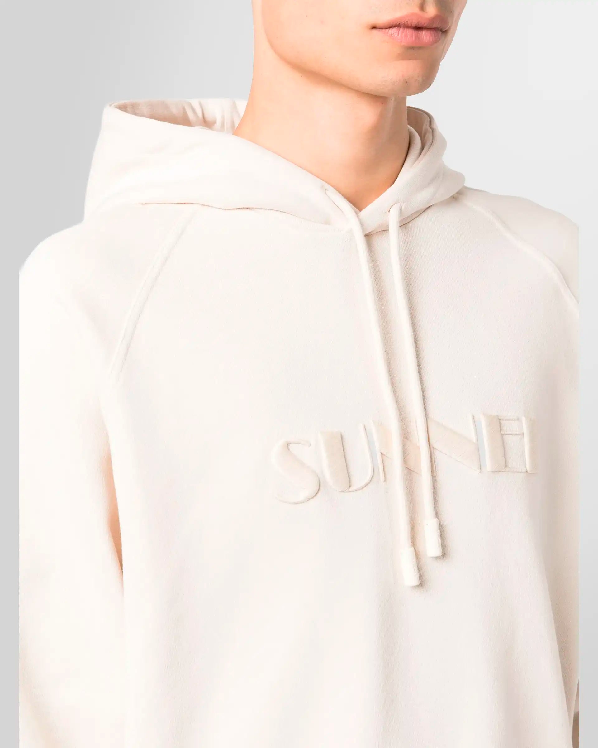 A close-up of the large, tonal embroidered Sunnei logo on the front of the light beige hoodie.