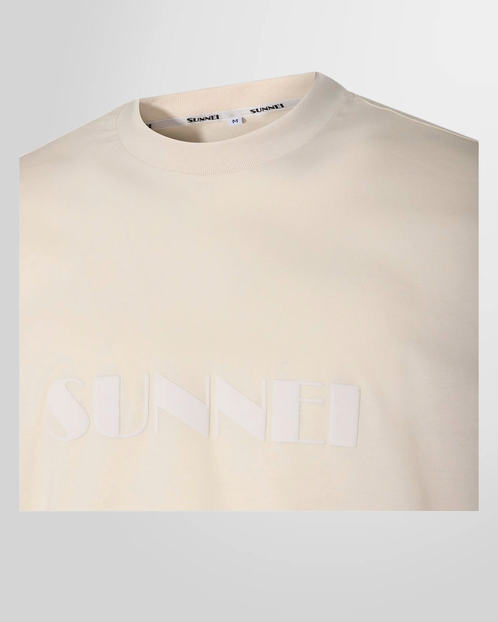 A close-up of the signature Sunnei logo printed on the front of the light beige t-shirt.