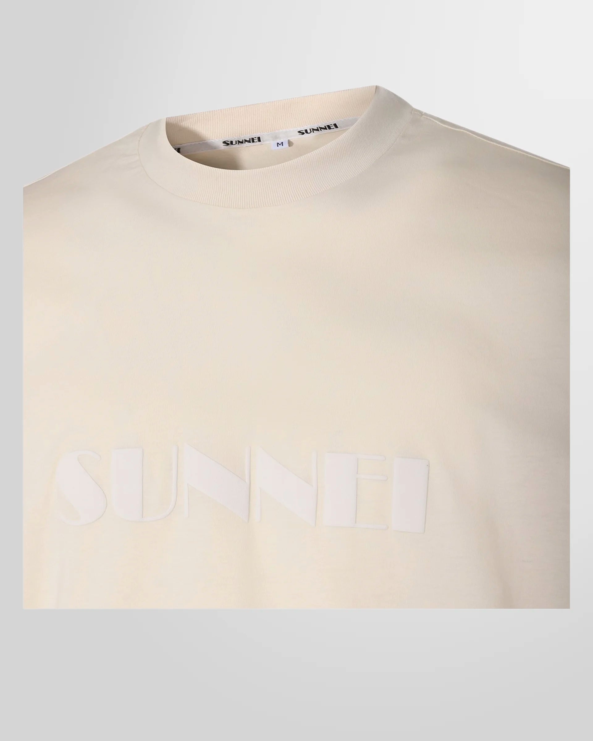 A close-up of the signature Sunnei logo printed on the front of the light beige t-shirt.